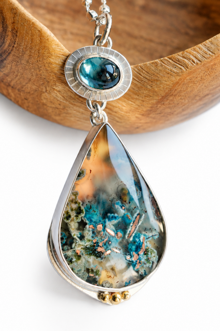A silver teardrop pendant necklace featuring a colorful geode-like inlay with blue, green, and orange tones, displayed on a white background with a wooden bowl prop.
