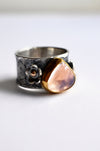 A silver ring with a wide band featuring floral engravings and a large, teardrop-shaped peach-pink stone accented by a gold bezel setting.
