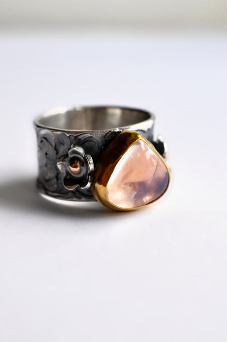 A silver ring with a wide band featuring floral engravings and a large, teardrop-shaped peach-pink stone accented by a gold bezel setting.